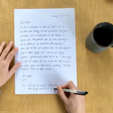 Eine Person schreibt mit einem Stift auf ein weißes Blatt Papier, welches auf einen Holztisch liegt