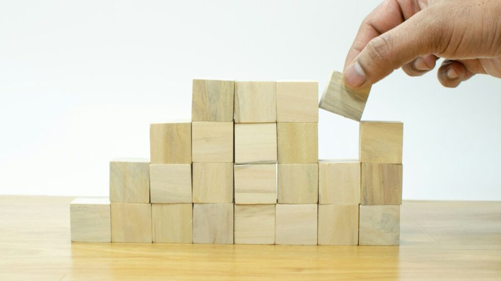 Zu sehen sind Bausteine oder Bauklötze, die aufeinander gestapelt sind. Eine Hand ergänzt einen in einer Lücke.