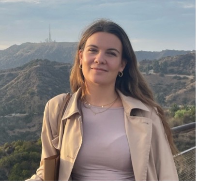 Lea hat lange hellbraune Haare, trägt eine beige Jacke und steht vor einer hügeligen Landschaft.