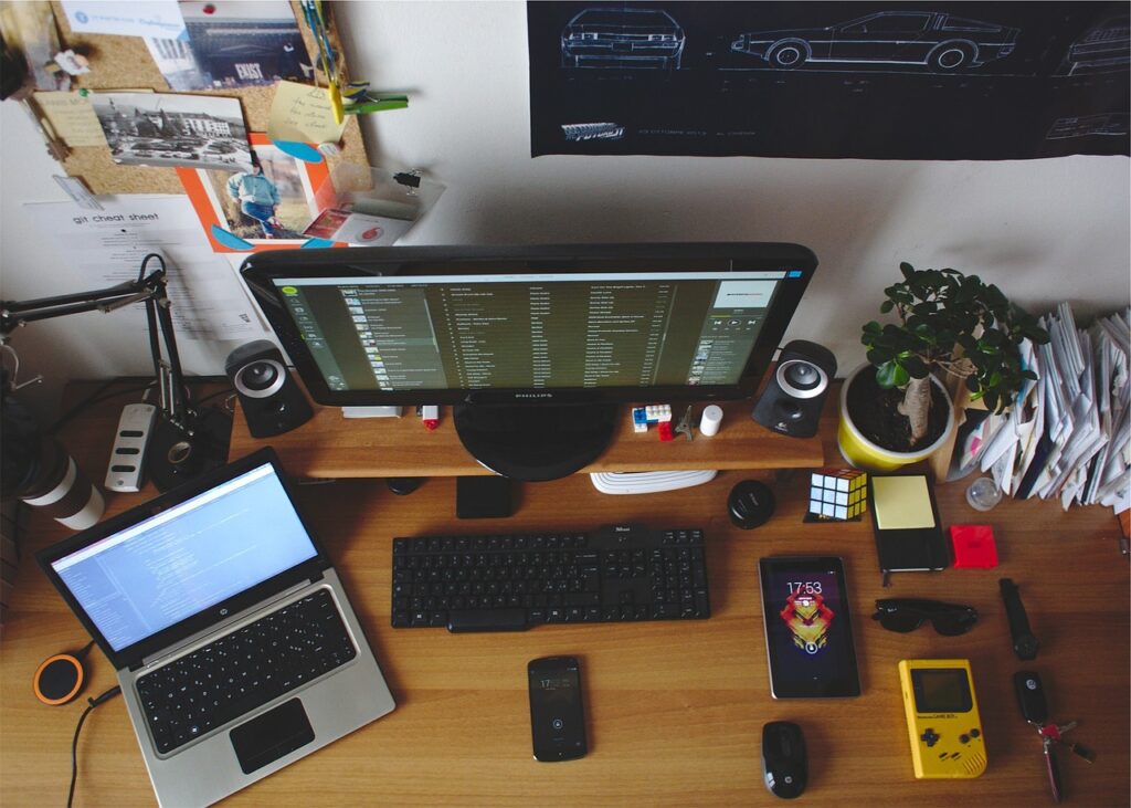 Auf dem Bild ist ein Schreibtisch zu sehen, auf dem ein Monitor und ein Laptop steht. Es soll eine Programmier-Szene darstellen.