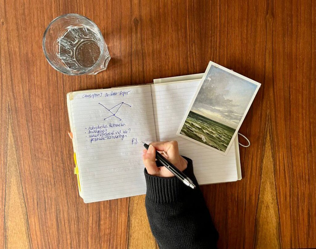Ein Schreibblock liegt auf einem Holztisch und jemand schreibt etwas mit einem Kugelschreiber hinein. Zu sehen sind noch ein Gals Wasser und eine Postkarte mit einer Zeichnung vom Meer
