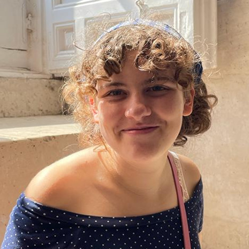Adriana hat kurze Locken, trägt ein Haarband und ein weiß gepunktetes Oberteil. Sie steht in einem beigen Raum vor einem Fenster.