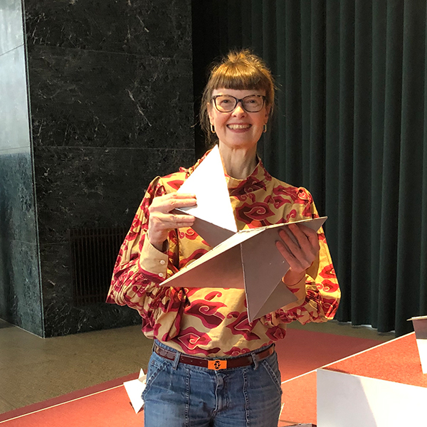 Portät von Astrid Mania, sie trägt eine bunte Bluse und hält ein graues Kunstobjekt in der Hand.