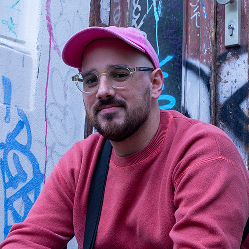 Augustin hat einen kurzen Bart und trägt eine Brille und eine rosa Cap.  Er sitzt vor einer Wand mit Graffiti.