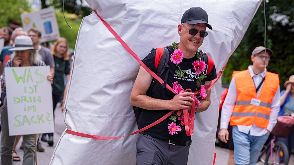Axel trägt Blumenschmuck um den Hals, wie eine Kuh beim Almabtrieb und auf dem Rücken einen Sack voller Gas.