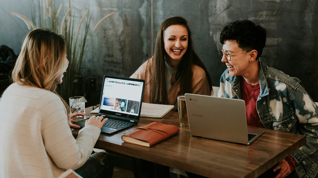 Drei lachende Frauen sitzen mit ihren Laptops zusammen.