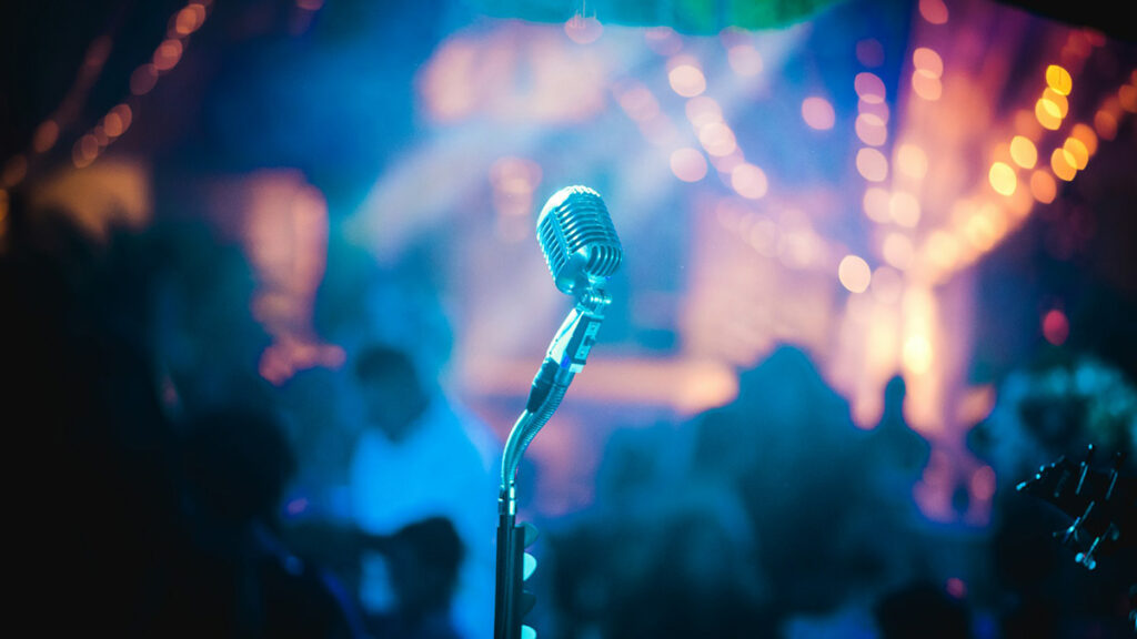 Auf dem Bild ist ein Mikrofon auf einer Bühne abgebildet. im Hintergrund leuchten bunte Lichter.