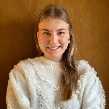 Elisabeth steht vor einer Holzwand, hat lange glatte Haare und trägt einen weißen Strickpullover.
