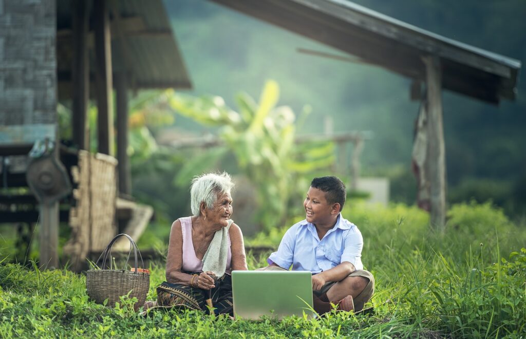 Eine ältere Frau sitzt in der Natur mit einem Jungen vor einem Laptop. Beide lachen und lernen voneinander.