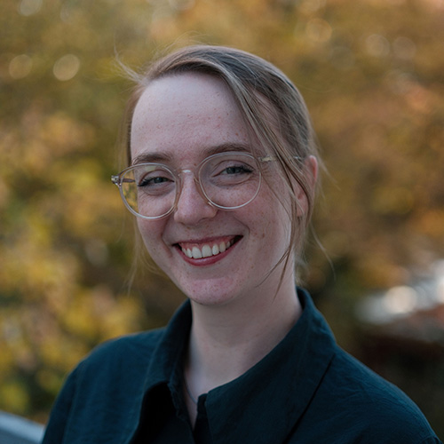 Marie trägt eine Brille mit durchsichtigem Gestell, einen Zopf und eine dunkelblaue Bluse.