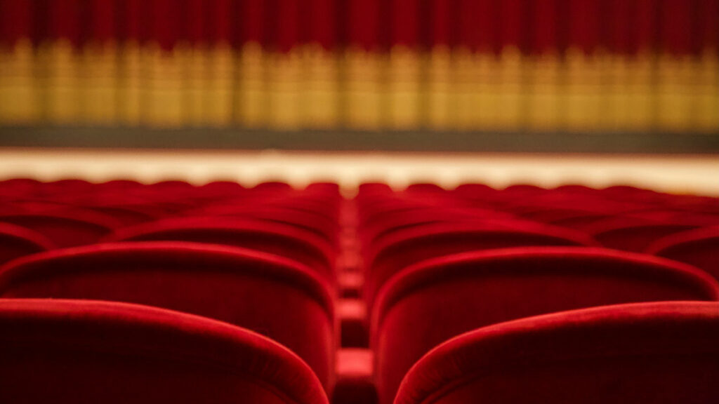 Das Bild zeigt die Rückenlehnen roter Theaterstühle, im Hintergrund ist der rote Vorhang der Bühne mit goldener Bordüre zu sehen.