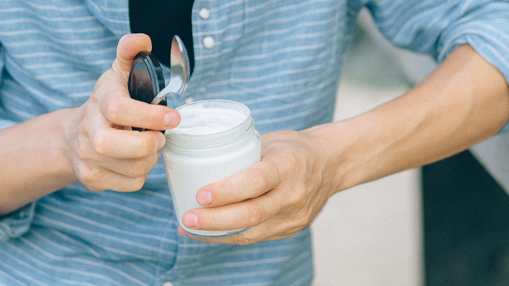 Eine Person öffnet ein kleines Glas mit Naturjoghurt und hat einen Löffel in der Hand.